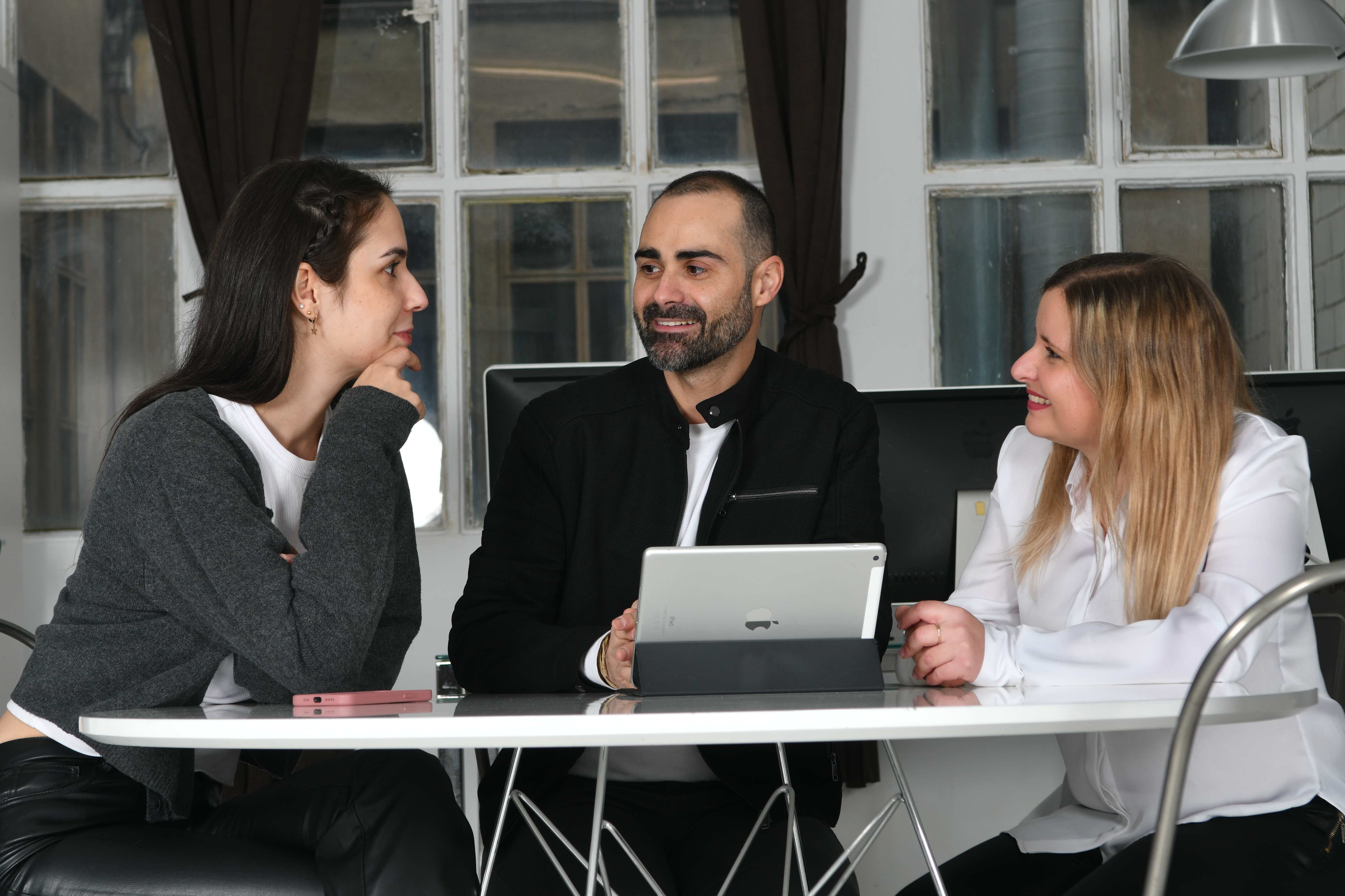 Tres profesionales de pie alrededor de una mesa revisando informes impresos y un portátil durante una sesión de trabajo.