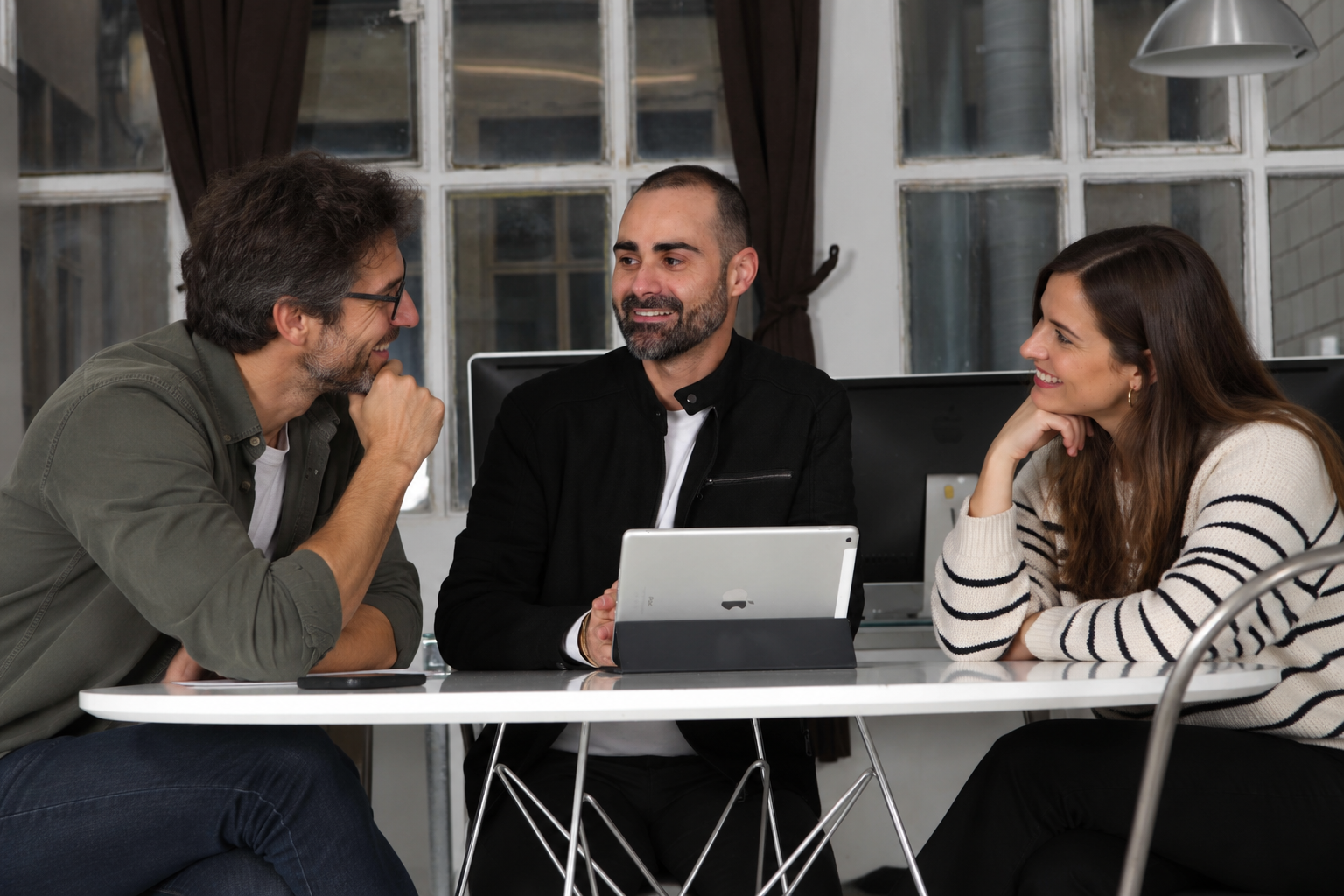 Tres profesionales de pie alrededor de una mesa revisando informes impresos y un portátil durante una sesión de trabajo.
